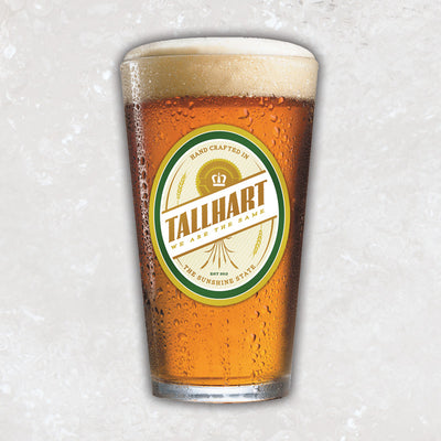 A pint glass with a oval shaped oat colored emblem. On the emblem it says  "TALLHART" in diagonal lettering. There is also text on the top and bottom that says "HAND CRAFTED IN" "THE SUNSHINE STATE".